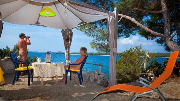 Para na plaży, relaksująca się w namiocie. Mężczyzna obserwuje morze przez lornetkę. To idealne miejsce na naturystyczny camping.