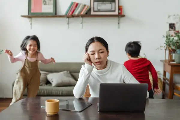 Donna esausta davanti al laptop, mentre i bambini giocano. Un quadro di **parental burnout** in casa.