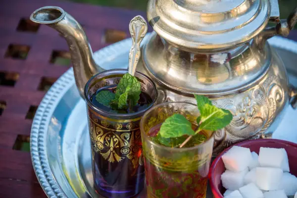 Zwei Gläser mit Minztee, garniert mit frischen Minzblättern, stehen neben einem traditionellen Teekessel und Zuckerwürfeln. Verschiedene Minze Arten verfeinern das Getränk.