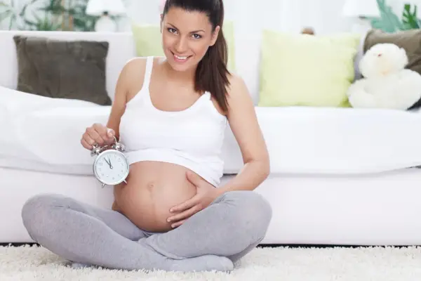 Uśmiechnięta kobieta w 40 tygodniu ciąży trzyma budzik. Czas na poród!