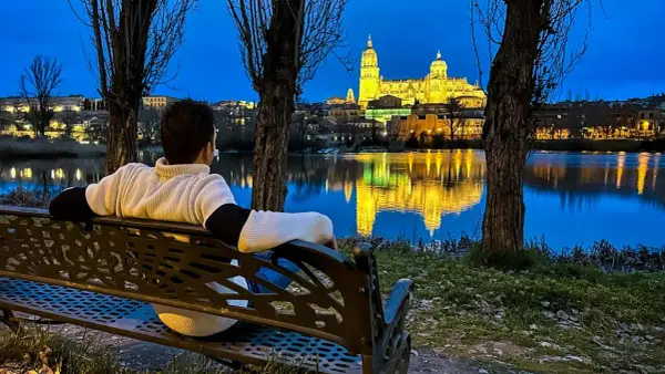 Persona sentada en un banco contempla la Catedral de Salamanca iluminada al anochecer, reflejada en el río. Ideal para saber qué ver en Salamanca en un día.