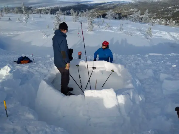 Två personer bygger en bivack i snön. De använder snöblock för att bygga väggarna och stavar för att markera utrymmet.