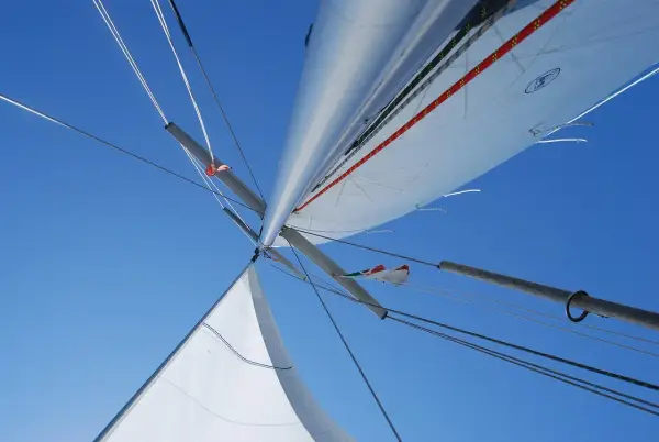 Vue plongeante sur le mat de bateau, voiles blanches déployées sous un ciel bleu éclatant, cordages tendus.