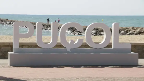Letras gigantes "PUZOL" en la playa, con el mar azul y personas al fondo.