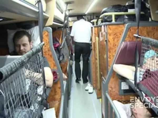 Intérieur d'un bus avec des couchettes. Un homme en blanc marche dans l'allée, tandis que des passagers se reposent dans leurs couchettes.