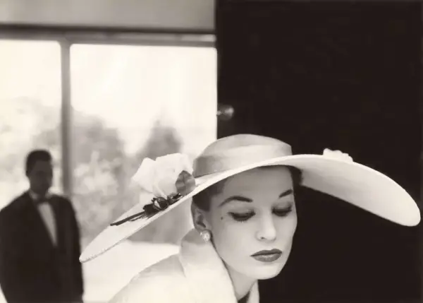 Mujer con sombrero de ala ancha y rosa, estilo Helmut Newton. Un hombre en traje con pajarita al fondo.