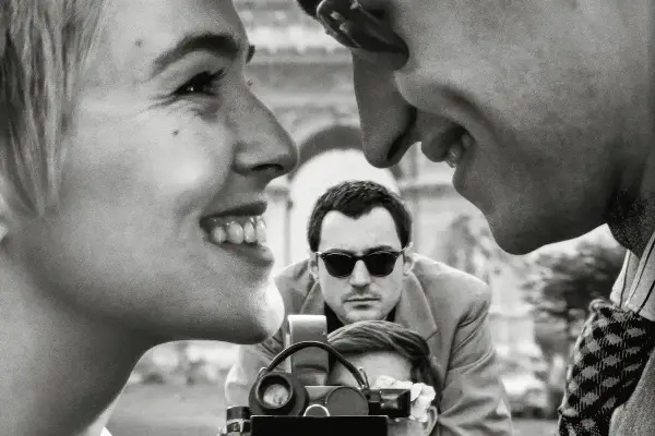Primo piano di una donna sorridente, con un uomo che la guarda e un altro uomo con occhiali da sole che filma. Un omaggio ai registi importanti.