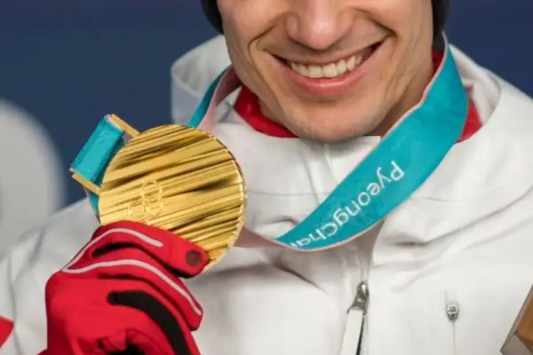 Uśmiechnięty sportowiec w czerwonej rękawicy trzyma złoty medal olimpijski z PyeongChang. Taki medal to marzenie o emeryturze.