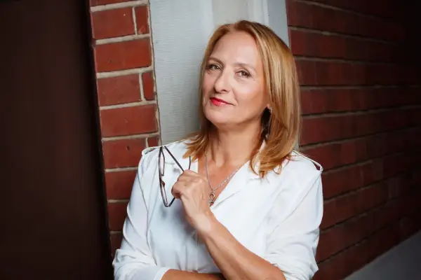 Felicitas Fuchs, eine Frau mit blondem Haar und rotem Lippenstift, hält eine Brille in der Hand. Sie steht vor einer Ziegelwand.