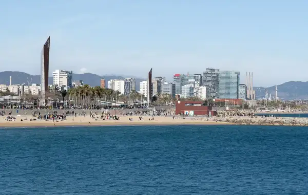 Ludzie odpoczywają na plaży, a w tle widać nowoczesne budynki Barcelony. To miasto leży nad Morzem Śródziemnym.