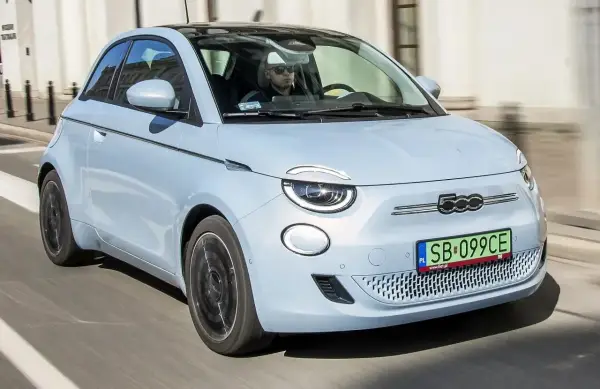 Jasnoniebieski fiat elektryczny 500 z czarnym dachem jedzie ulicą. Kierowca w okularach przeciwsłonecznych prowadzi auto.