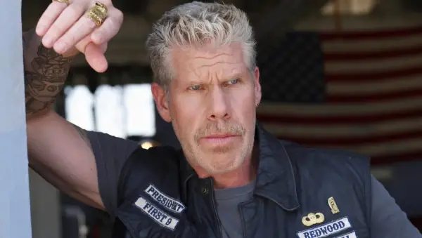 Ron Perlman w skórzanej kamizelce z naszywkami, z tatuażem na ramieniu i pierścieniami na palcach, na tle amerykańskiej flagi.