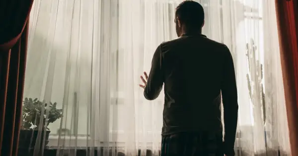 Mężczyzna stoi przy oknie, zasłoniętym przezroczystą tkaniną. Jego dłoń opiera się o materiał, jakby szukał kontaktu ze światem zewnętrznym. Obraz symbolizuje schizofrenia objawy, izolację i trudności w komunikacji.