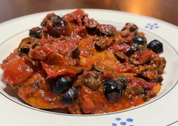 Bistecche alla pizzaiola, un piatto succulento con pomodoro, olive nere e capperi, servito su un piatto bianco con decorazioni blu.