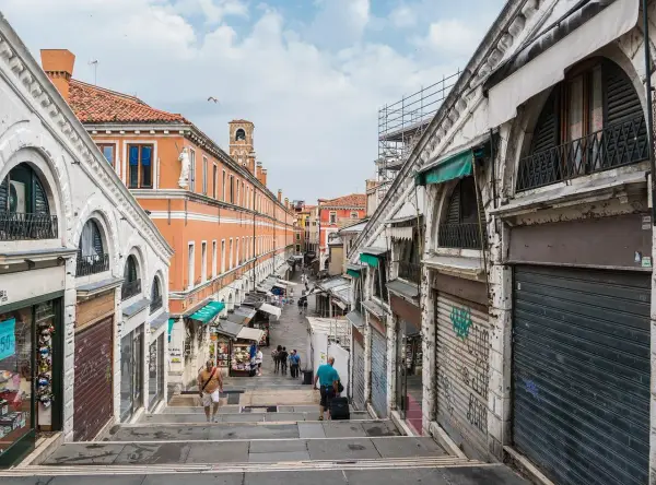 Scendendo le scale del ponte di Rialto a Venezia, si intravede un mercato vivace con negozi e bancarelle.