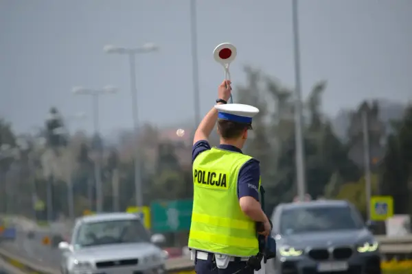 Policjant kieruje ruchem, podnosząc znak stop. W tle widać samochody i słupy oświetleniowe, sugerujące podwyższone **stopnie alarmowe**.