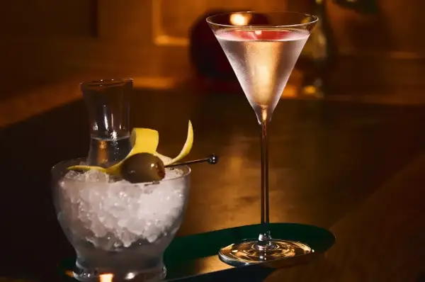 Elegancki drink z martini w kieliszku, obok miseczka z lodem, oliwką i skórką cytryny.