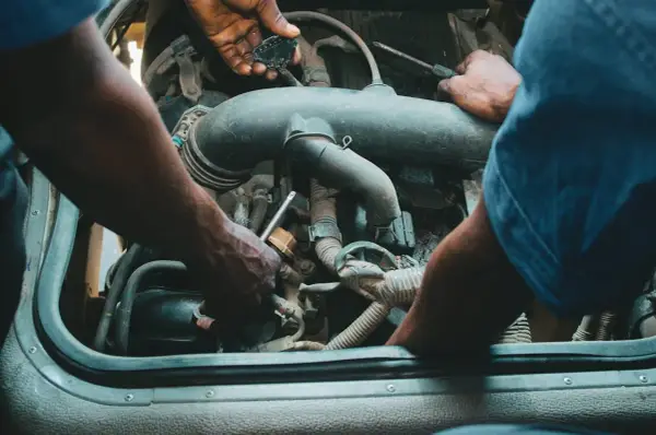 Mechanicy pracują przy silniku, jeden z nich trzyma wtyczkę, drugi dokręca śrubę. Może to być naprawa zaworu par paliwa.