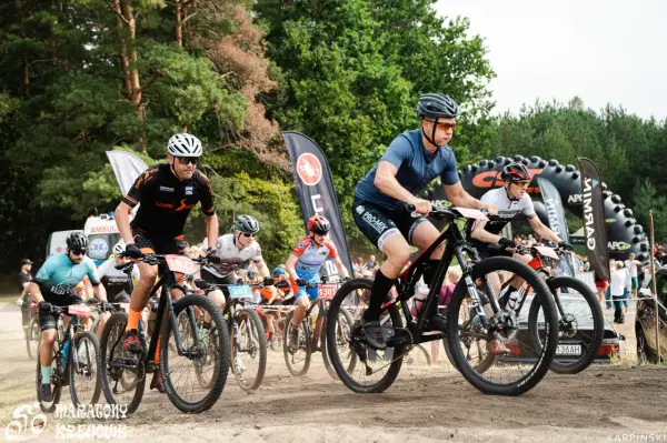 Kolarze biorą udział w wyścigu **zawody MTB**. Widać ich w akcji, jadących po piaszczystej trasie, z lasem w tle.