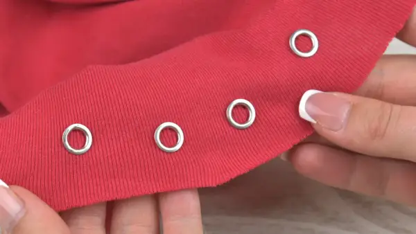 Demonstration of how to put metal rings in fabric. A hand holds red fabric with several silver grommets already attached, and another grommet is about to be placed.