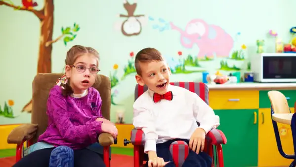Dwoje dzieci w fotelikach, dziewczynka w okularach i chłopiec w muszce, na tle kolorowej ściany z rysunkami.