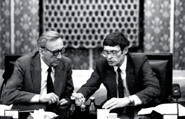Dwóch mężczyzn w garniturach przy stole, dyskutujących o historii politycznej Polski. Antoni Dudek analizuje dokumenty.