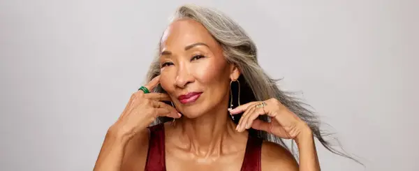 Mujer con cabello gris, con un elegante efecto de grey blending, sonríe con labios rojos y joyas doradas y verdes.