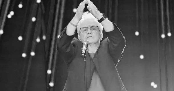 Starsza pani z białymi włosami, w okularach, z rękami uniesionymi nad głową, przed mikrofonem. To może być Jolka podczas występu.