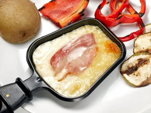 Un poêlon de raclette avec fromage fondu et lardons, accompagné d'une pomme de terre, de poivrons rouges grillés et de courgettes. Une recette de sauce raclette facile et gourmande.