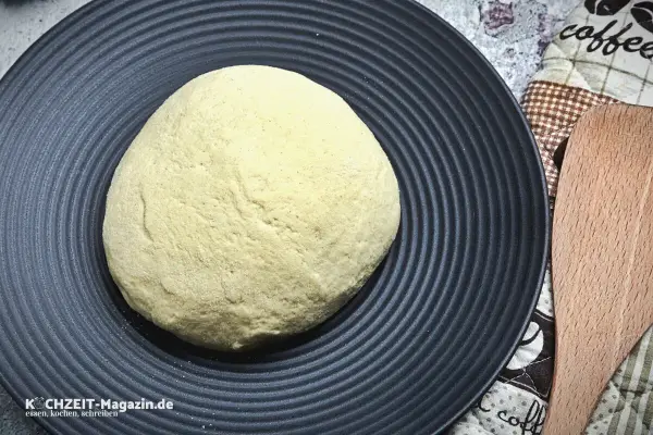 Ein runder, heller pizzateig wie beim Italiener liegt auf einem schwarzen Teller. Daneben ein Holzspatel und eine Kaffeetasse-Serviette.