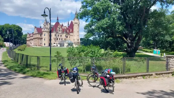 Trzy rowery z sakwami czekają przed zamkiem. W tle rozciąga się zielony park, a w oddali widać fragment rzeki Niemen.