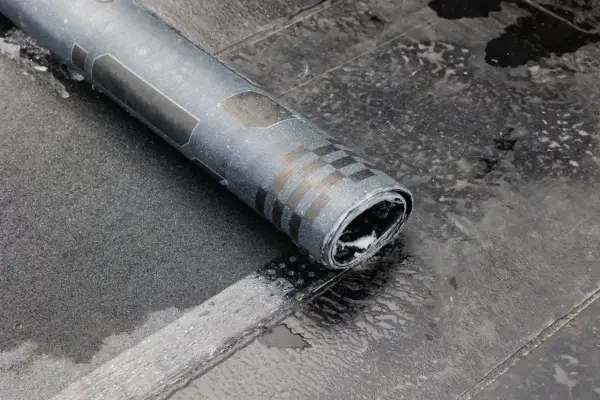 Rolka papy na zimno leży na mokrym dachu. Widać fragmenty wzoru na papie i jej zwinięty koniec.