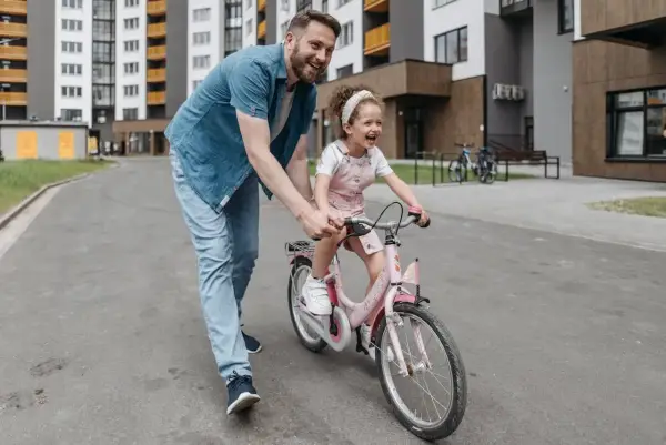Tata uczy córkę, jak nauczyć dziecko jazdy na rowerze. Radosna dziewczynka na różowym rowerze, tata ją asekuruje.