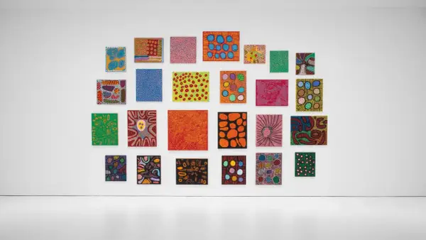 A vibrant collection of paintings by top visual artists, featuring bold colors and abstract patterns, adorns a white gallery wall.