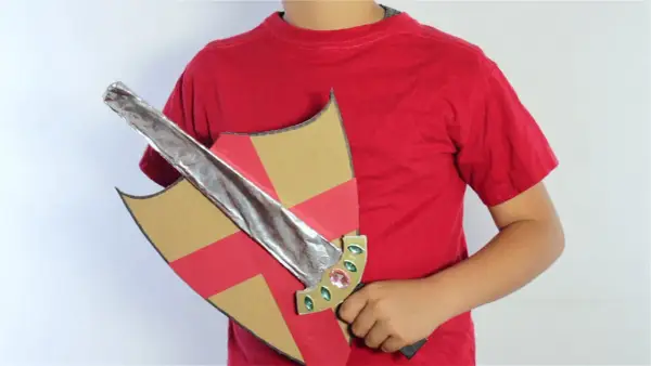 Un niño con camiseta roja sostiene un escudo de cartón con detalles rojos y dorados y una espada de cartón plateada, lista para la aventura.