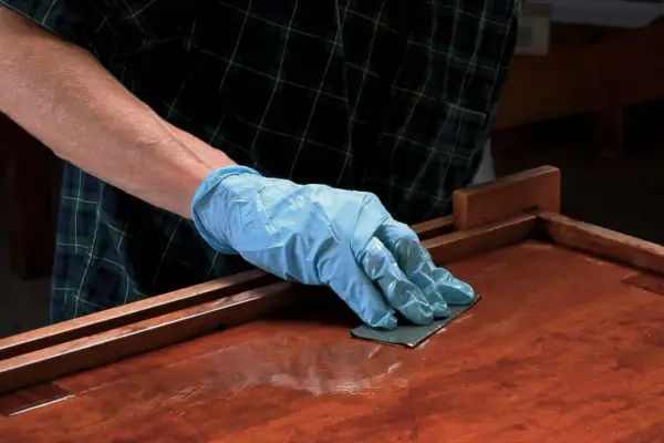 Mano enguantada lijando superficie de madera barnizada. El lijado es un paso clave para aplicar lac.