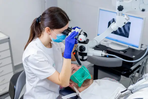 Dentystka w masce i rękawiczkach bada ząb pacjenta pod mikroskopem. Na ekranie widać zdjęcie rentgenowskie. To nowoczesne leczenie kanałowe, cena zabiegu zależy od skomplikowania.