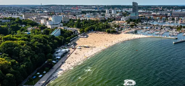 Słoneczny bulwar nadmorski Gdynia tętni życiem. Plaża pełna ludzi, a w oddali port z żaglówkami i miejska panorama.