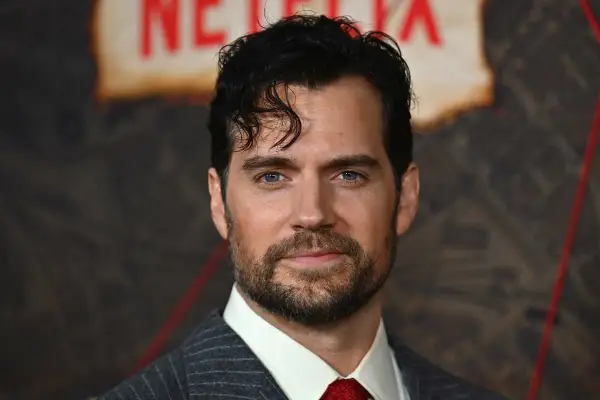 Henry Cavill w garniturze i czerwonym krawacie, z zarostem i niebieskimi oczami.
