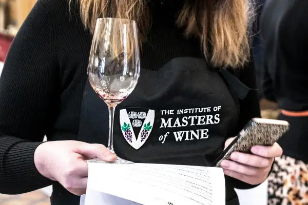 Eine Person mit einer Schürze des "Institute of Masters of Wine" hält ein Weinglas und ein Smartphone.