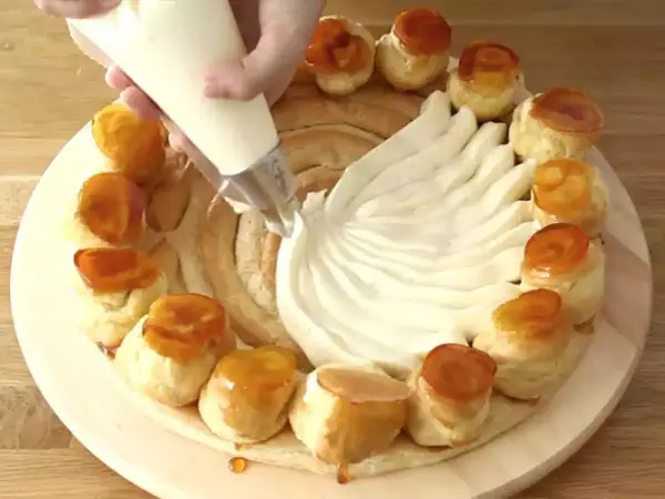 Une main garnit une couronne de choux avec de la crème chiboust, créant un dessert spectaculaire.