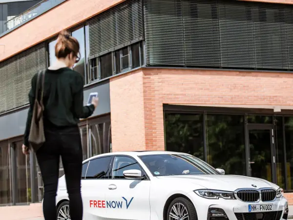 Frau mit Smartphone wartet auf FREE NOW Fahrt. Keine Beschwerde, nur Vorfreude auf die Fahrt.