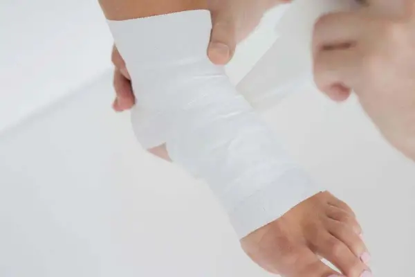 Dłonie owijają stopę białą taśmą do tapingu, zapewniając wsparcie i stabilizację.