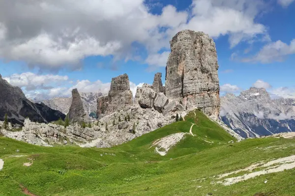Sentiero che serpeggia tra verdi prati verso le maestose Cinque Torri Dolomiti, sotto un cielo azzurro con nuvole bianche.