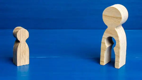 Zwei Holzfiguren auf blauem Grund. Eine kleine Figur steht allein. Die größere Figur hat eine leere Öffnung, die an ein Kind erinnert, und symbolisiert das Bedauern über Mutterschaft.