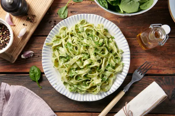 Pyszne zielone tagliatelle z pesto, podane na talerzu z niebieskimi paskami. Obok garnek z pieprzem, czosnek i oliwa.