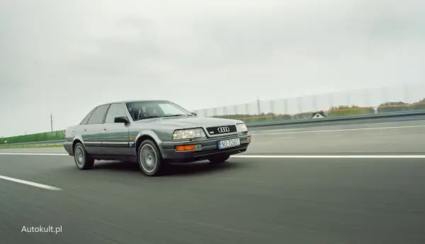 Szary Audi V8 pędzi po autostradzie, chmury na niebie i zielone drzewa w tle.