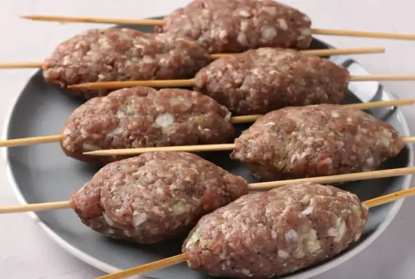 Siedem surowych kofta kebab na patyczkach, ułożonych na szarym talerzu, gotowych do grillowania.