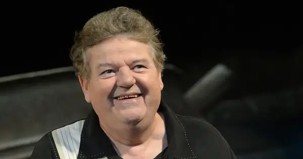 Uśmiechnięty Robbie Coltrane, który grał Hagrida, w czarnej koszuli z białymi pasami.