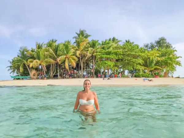Mujer sonríe en aguas cristalinas, con una isla tropical llena de palmeras al fondo. ¡Un sueño para viajar a Filipinas!
